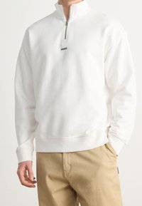 Felpa bianca con collo alto e mezza zip, caratterizzata da un piccolo logo nero "HUGO" sul petto. Materiale in morbido misto cotone.