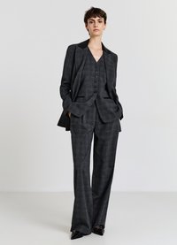 Grau karierter Anzug bestehend aus einem maßgeschneiderten Blazer, einer Weste mit Knöpfen und weiten Hosen; schwarze Akzente am Kragen des Blazers.