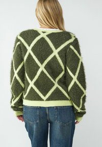 Zielony puszysty sweter z diamentowym wzorem w jasnym żółtym kolorze. Żebrowany dół i mankiety, o luźnym kroju. Noszony z niebieskimi dżinsami.