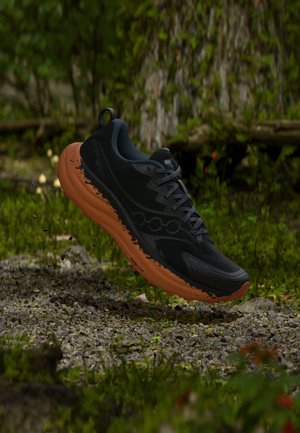 Schwarzer und orangefarbener Trailrunning-Schuh mit Schlammspritzern, schwebend über einem felsigen Waldboden mit grünem Laub im Hintergrund.