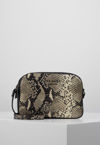 Malá obdélníková crossbody taška s černým a béžovým hadím vzorem a tenkým nastavitelným popruhem, umístěná na bílé podložce.