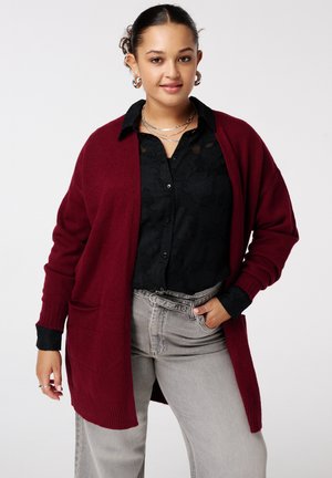 Femme portant une chemise noire à motif, un cardigan bordeaux ouvert, un jean gris à taille haute, des boucles d'oreilles argentées en anneau et des colliers superposés.