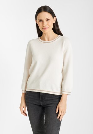 Vrouw met lang zwart haar die een witte gebreide trui met beige rand en donkergrijze spijkerbroek draagt, staand tegen een eenvoudige witte achtergrond.