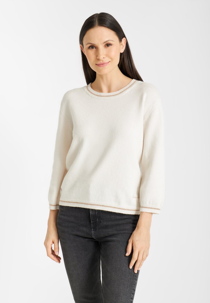 Vrouw met lang zwart haar die een witte gebreide trui met beige rand en donkergrijze spijkerbroek draagt, staand tegen een eenvoudige witte achtergrond.