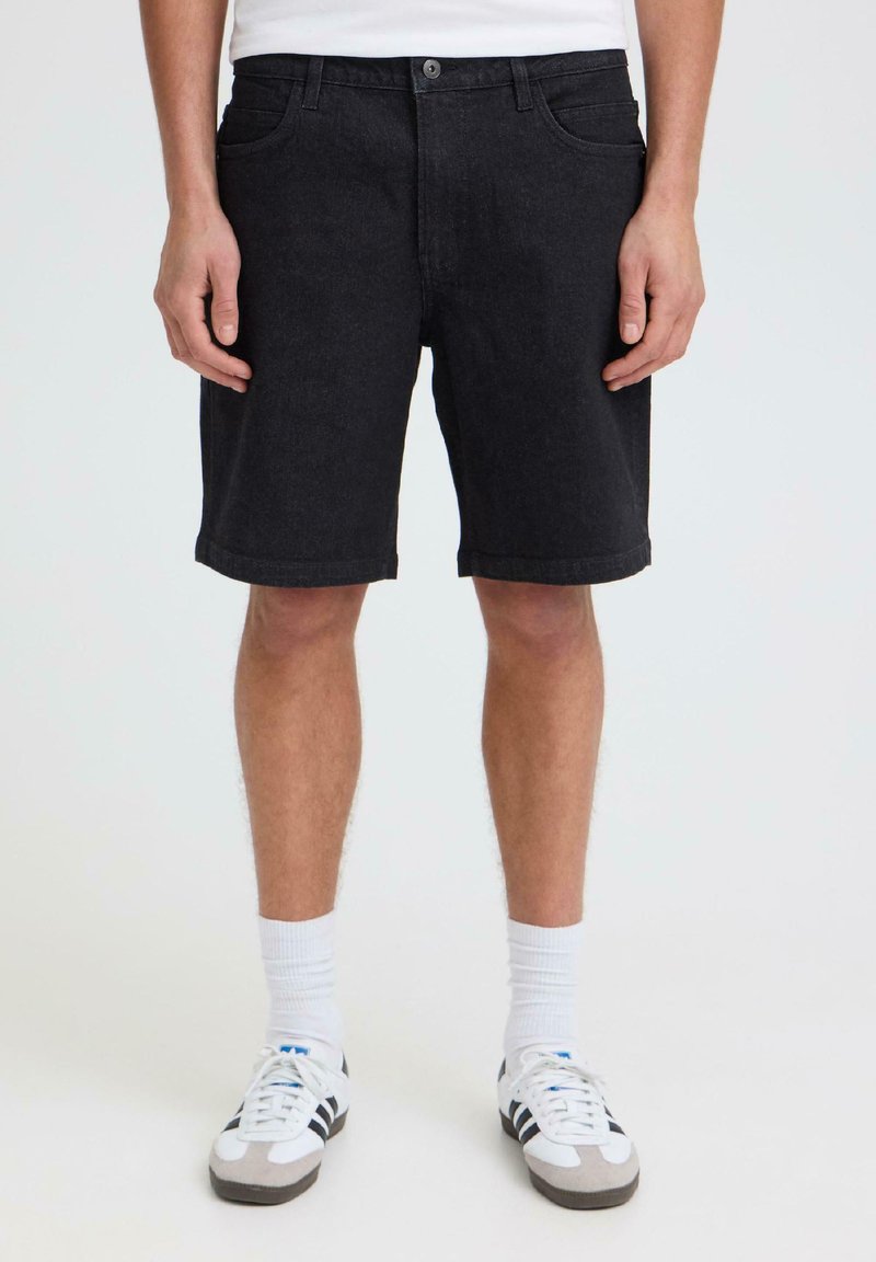 Homme portant un short en jean noir, des chaussettes blanches montantes et des baskets blanches à rayures noires, debout devant un fond clair uni.