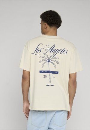 Mann von hinten, trägt ein beiges T-Shirt mit einer blauen Palme und dem Text "Los Angeles Urban Classics 2015" auf dem Rücken, kombiniert mit hellblauen Jeans.