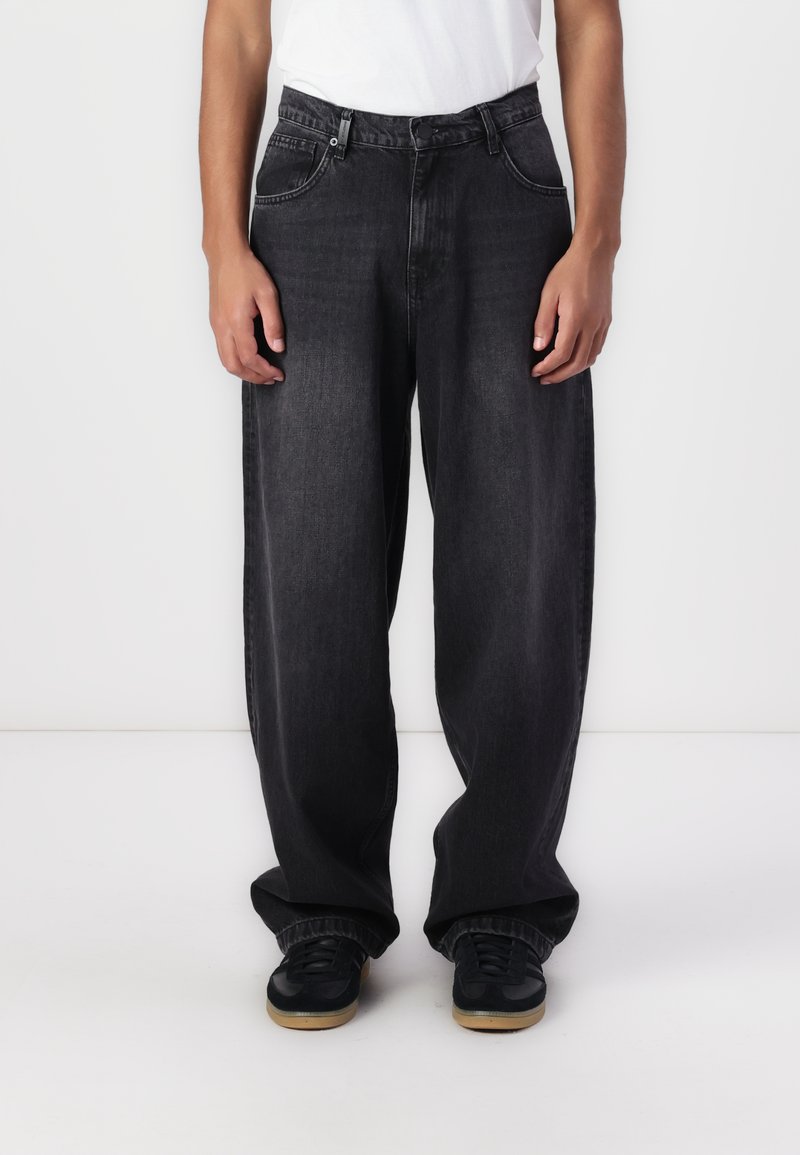 Jeans negri cu croi larg, din material denim, cinci buzunare, bucle pentru curea, decolorare subtilă, asortați cu teniși negri pe un fundal deschis.