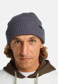 Berretto lavorato a maglia grigio con una texture a coste e un piccolo logo nero come dettaglio. Design semplice e arrotondato che si adatta perfettamente alla testa.