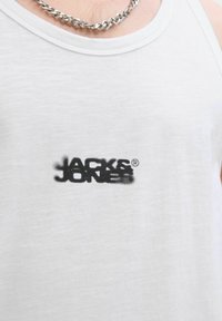 Bílý bavlněný tank top s černým, částečně vybledlým logem "JACK & JONES" na přední straně, s kulatým výstřihem a jemnou texturou.