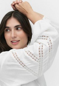 Weiße Bluse mit langen Ärmeln, die mit kunstvollen Spitzenausschnitten an den Ärmeln versehen ist. Glatte Textur, lockere Passform und subtiler Glanz.
