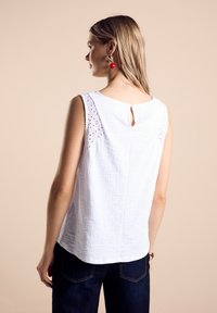 Witte mouwloze top met geborduurde kantaccenten op de schouders, textuurstof en een keyhole/knopen sluiting aan de achterkant. Draagt met donkere jeans.