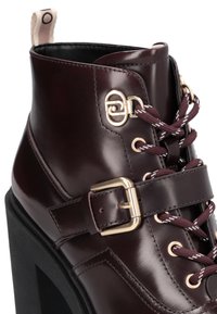 Burgunderfarbene Leder-Stiefelette mit einem klobigen schwarzen Absatz, goldenen Beschlägen, dekorativer Schnalle und kastanienbraunen sowie weißen Schnürsenkeln.