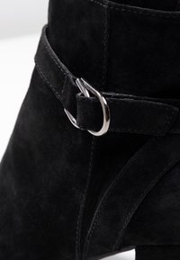 Botte de cheville en daim noir avec une large sangle et un détail de boucle en argent, offrant une silhouette élégante. Texturé doux avec des coutures raffinées.