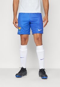 Blå fotbollsshorts med vertikala ränder och broderad tupp, tillsammans med vita strumpor och svarta fotbollsskor med blå detaljer. Nike-logotypen är närvarande.