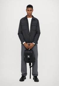 Black padded jacket with two chest pockets, grey textured pants, and a black leather backpack featuring a silver clasp and adjustable strap.