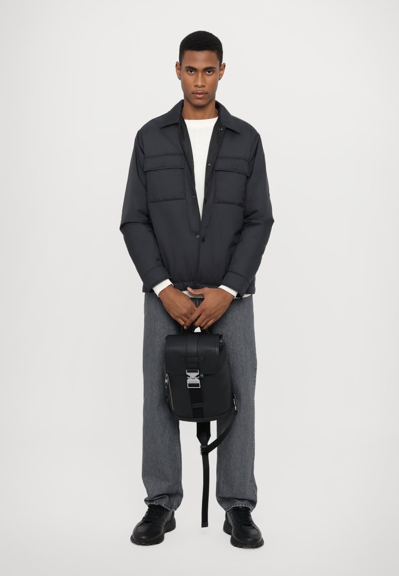 Black padded jacket with two chest pockets, grey textured pants, and a black leather backpack featuring a silver clasp and adjustable strap.