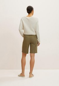 Haaktextuur, lichtbeige trui met een brede halslijn, gecombineerd met olijfgroene shorts met twee achterzakken. Sandalen maken de look compleet.