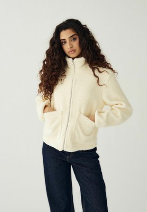 Mujer con cabello largo y rizado, vestida con una chaqueta polar cremosa con cremallera y bolsillos, y jeans oscuros, de pie frente a un fondo liso.