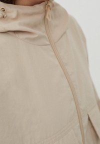 Chaqueta beige hecha de tejido ligero, con cierre frontal de cremallera, capucha ajustable y bolsillos laterales con detalles acolchados.