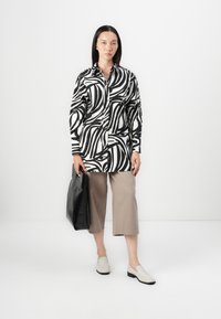 Camicia a righe in bianco e nero con colletto, abbinata a pantaloni beige a gamba larga e scarpe slip-on chiare. Tiene una borsa a tracolla in pelle nera.