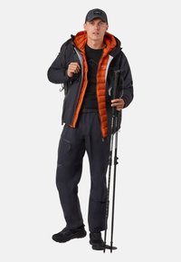 Zwarte buitenjas met oranje voering, gecombineerd met zwarte geïsoleerde broek. Draagt wandelstokken, met een rugzak en pet.