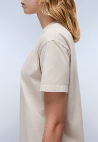Beige katoenen t-shirt met korte mouwen, voorzien van een ronde halslijn en een relaxte pasvorm, weergegeven vanaf de zijkant tegen een lichte achtergrond.