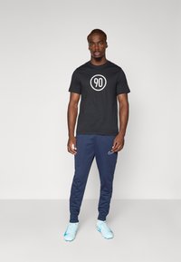 T-shirt en coton noir avec un graphique circulaire blanc portant le chiffre "90" au centre, associé à un pantalon bleu marine et des chaussures de sport bleu clair.