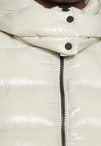 Nærbillede af en blank lyse beige pufferjakke med sort lynlås og trykknapper ved kraveområdet.