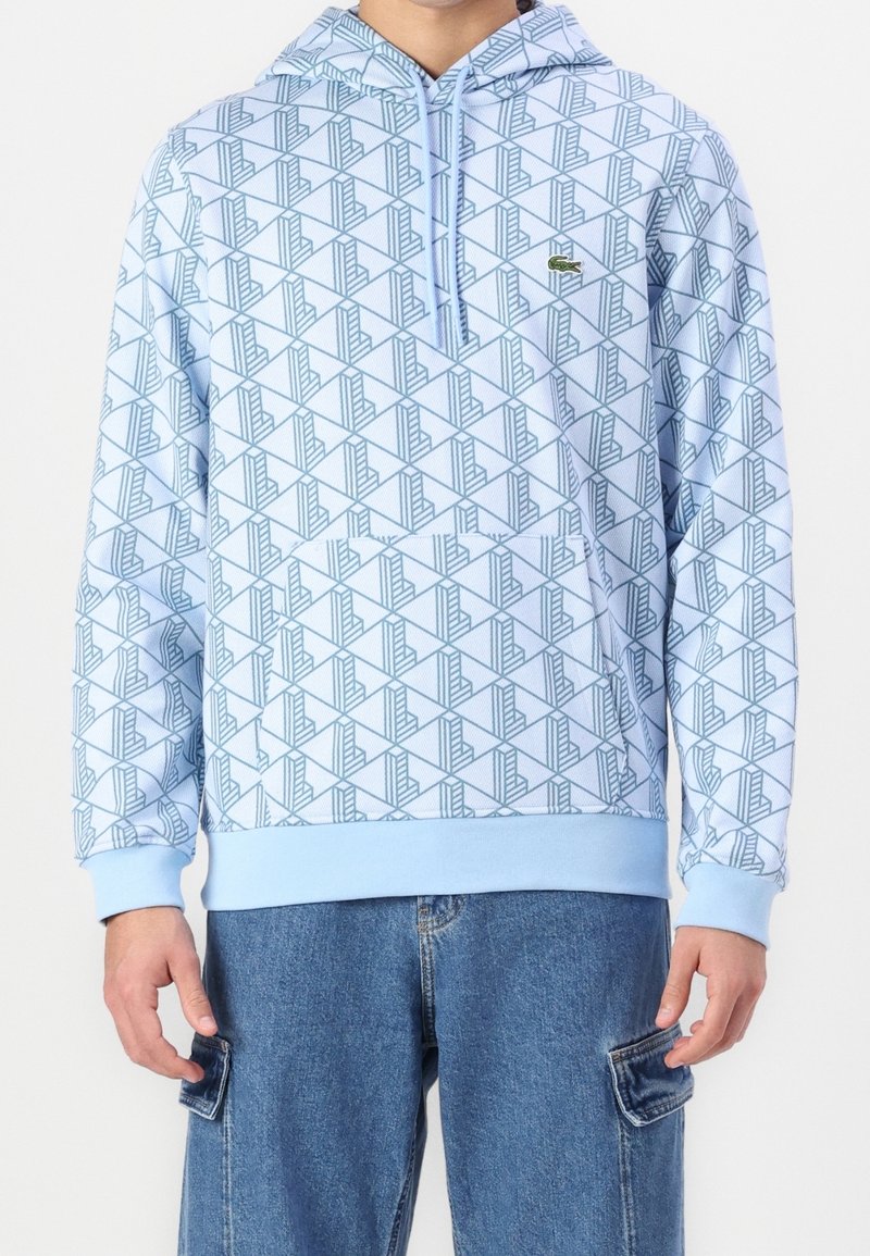Světle modrá mikina s kapucí s geometrickým trojúhelníkovým vzorem a malým zeleným logem krokodýla, nošena s modrými denimovými cargo kalhotami.