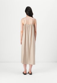 Bež maxi haljina s nabranym leđnim detaljem, tankim podesivim naramenicama i mekom, glatkom teksturom koja pada do visine sredine teleća. Vidljive crne sandale.