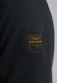 Črn waffle-pleten material z kvadratnim vezenim logotipom, ki vsebuje zlate črke in poudarke. Besedilo glasi: "PME LEGEND TESTED AT HIGH ALTITUDE."