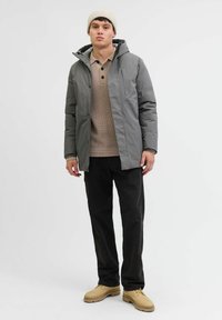 Grå dunjakke med hætte, beige striktrøje med knapper, sorte bukser og brune sko. Ensfarvet baggrund. Uformel lagdeling.