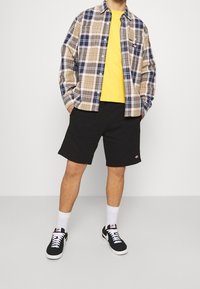 Kostkovaná košile na zapínání v béžových, tmavě modrých a oranžových odstínech, zkombinovaná se žlutým tričkem, černými šortkami a černými teniskami s bílými detaily.