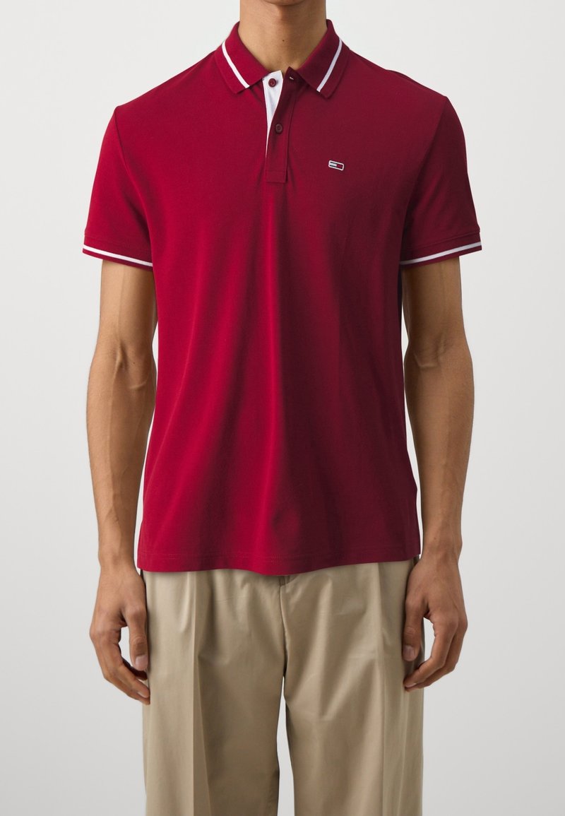 Homme portant un polo rouge à manches courtes avec des bordures blanches sur le col et les manches, associé à un pantalon beige sur un fond uni.