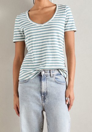 Person trägt ein kurzärmeliges, hellblau-weiß gestreiftes V-Ausschnitt-Shirt, das in eine hoch taillierte, hellblaue Jeans gesteckt ist, mit einem silbernen Ring an der rechten Hand.