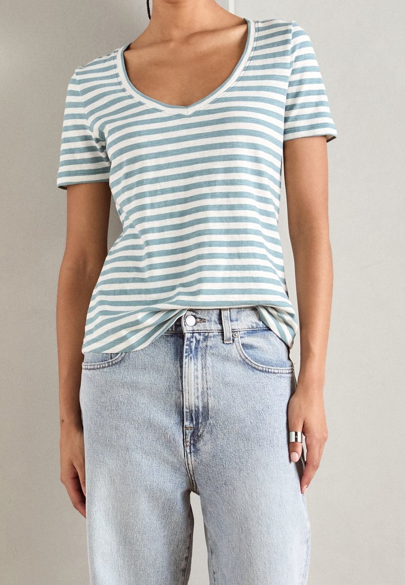 Personne portant une chemise à manches courtes, col en V rayée bleu clair et blanc, rentrée dans un jean taille haute délavé clair, avec une bague en argent à la main droite.