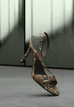 Πέδιλο με ψηλό τακούνι, καφέ και μαύρο σχέδιο φιδιού, με λουράκι στον αστράγαλο και αγκράφα, απεικονίζεται πάνω σε θολό πράσινο και γκρι φόντο.