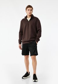 Brun zip-up tröja, svarta shorts och sneakers. Mjuk textur, regular fit. Tröjan har en ståkrage och ribbade ärmslut.