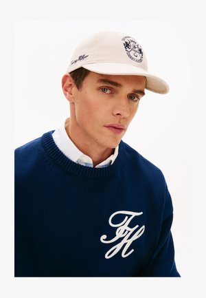 Jeune homme portant une casquette crème avec un logo et un pull bleu brodé des initiales blanches "TH" sur une chemise blanche boutonnée.