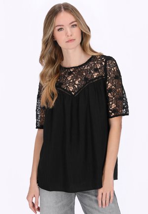 Ung kvinde med langt, bølget hår, iført en sort blonderbluse med korte ærmer og grå jeans, stående mod en ensartet hvid baggrund.