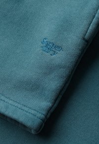 Nærbillede af turkis stof med foldet ærme med blåt broderet "Super Dry"-logo.