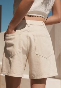 Shorts en denim beige avec une taille mi-haute, des poches arrière et une coupe droite, portés avec un crop top blanc.