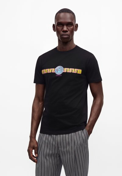 Ανδρας που φοράει μαύρο T-shirt Versace με πολύχρωμο λογότυπο Μέδουσα και γεωμετρικό μοτίβο, συνδυασμένο με γκρι και λευκό ριγέ παντελόνι.