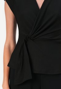 Haut noir enroulé en tissu lisse, avec un détail de nœud torsadé à la taille et des manches courtes, offrant une coupe décontractée.
