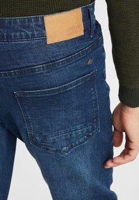 Homme portant un jean en denim bleu foncé et un pull en laine vert, vu de dos en se concentrant sur la zone de la poche droite.