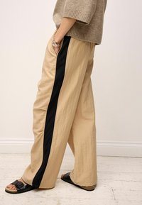 Breddebenede bukser i beige stof med en glat tekstur, udstyret med en sort sidelinie og parret med sorte sandaler.