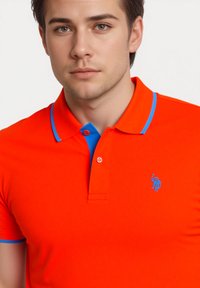 Červené polo tričko s modrým lemováním límečku, se dvěma knoflíky a vyšitým modrým logem na levé části hrudi. Hladká textura látky.