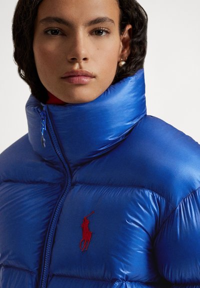 Blue puffer jacket with a high collar, featuring a red embroidered logo. Material appears shiny with quilted sections for insulation.