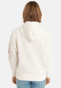Cream-colored hoodie with a relaxed fit, featuring a hood, ribbed cuffs, and floral embroidery on the left sleeve. Soft fabric texture.
