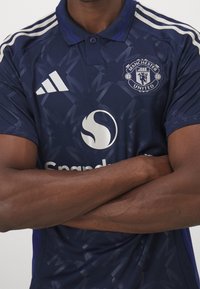 Granatowa koszulka polo Manchester United z delikatnym wzorem, białymi paskami Adidas i logo sponsora z przodu.